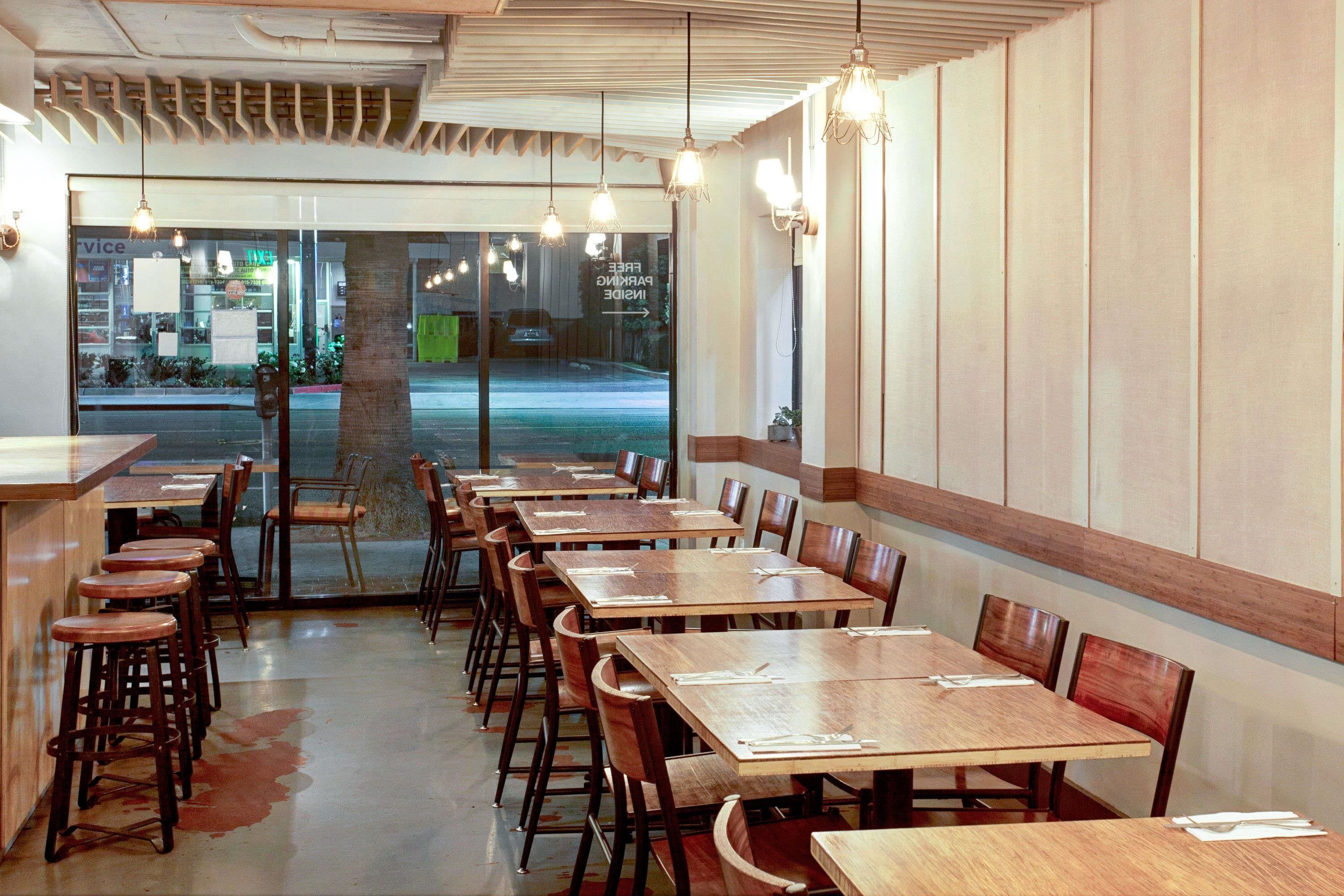 L’intérieur d’un restaurant présente de nombreuses tables et chaises en bois, un coin bar avec des tabourets et des lampes suspendues.