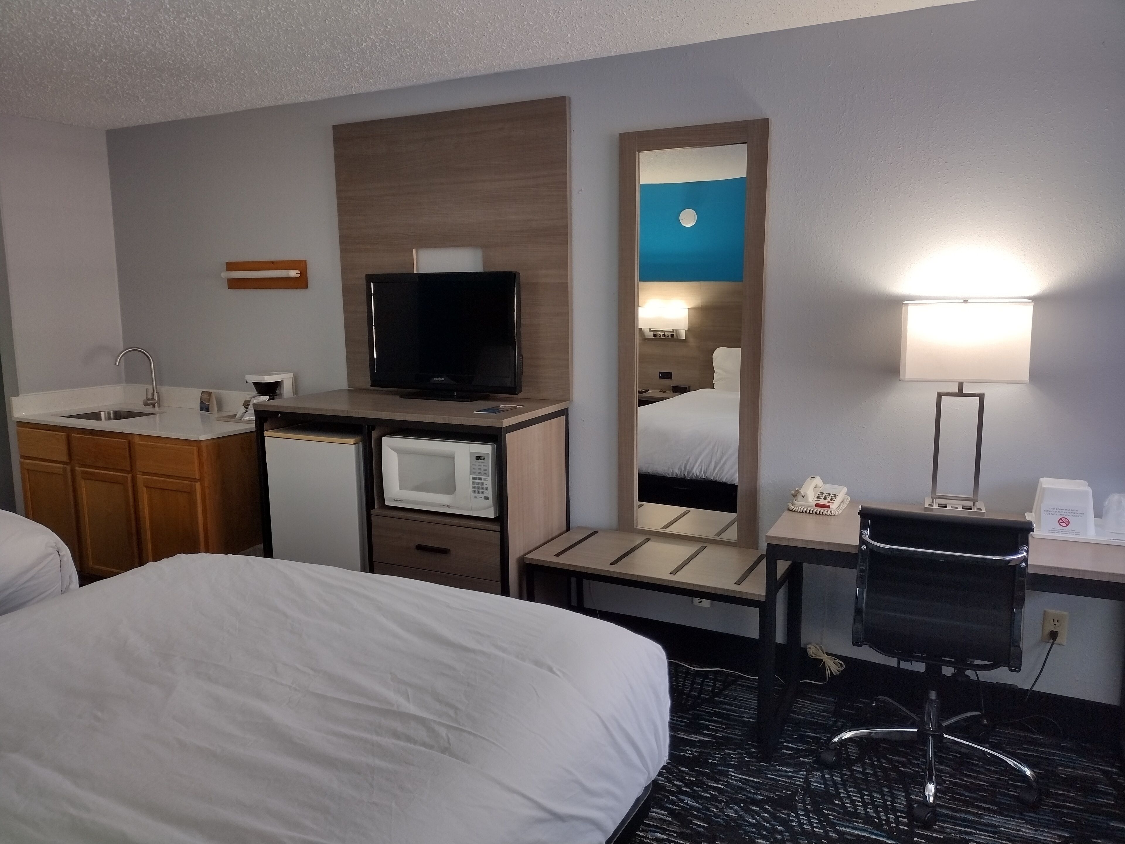 A hotel room with a bed, a desk with a lamp, a television, a microwave, a small refrigerator, and a mirror.
