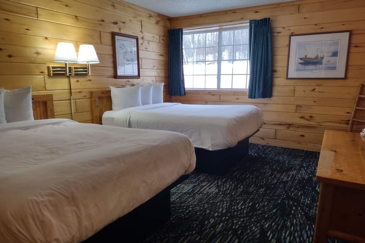 Ein blockhaushaltähnliches Zimmer mit zwei Betten, Holzboden, einem fenster mit Vorhängen und einer Lampe.