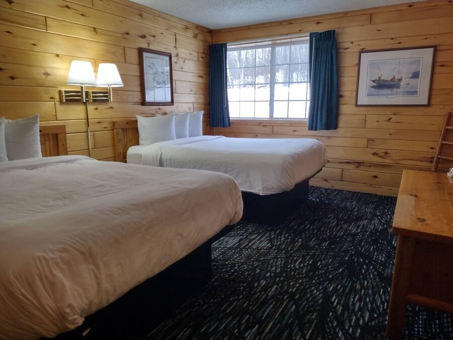 A cabin-style room with two beds, a wooden floor, a window with curtains, and a lamp.
