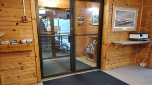 Ein Holzblockhaus-Interieur mit Glastür zu verschneiter Landschaft, eine kleine Ablage mit Snacks, ein gerahmtes Bild an der Wand und ein Schreibtisch mit einem Computer.