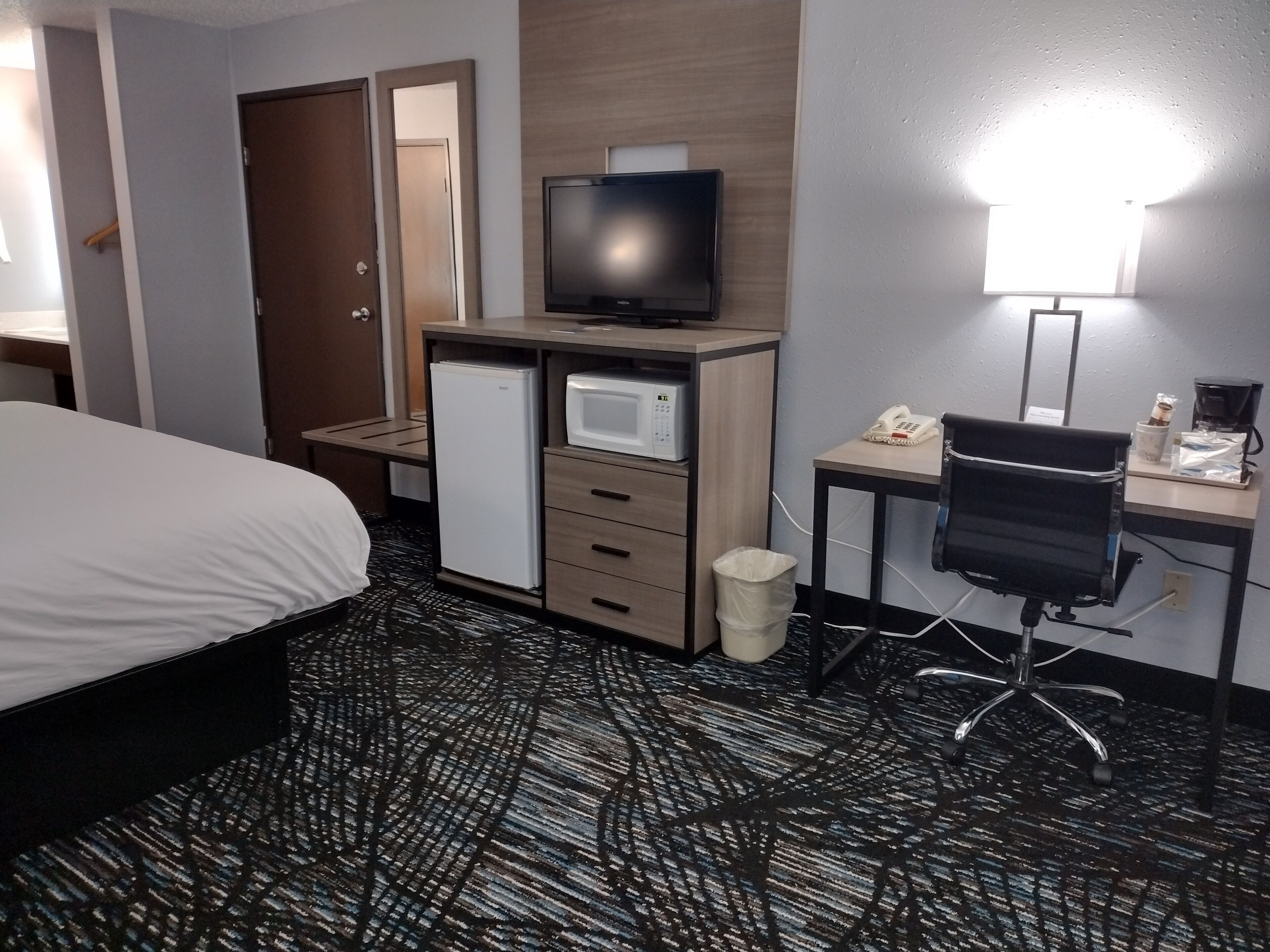 A hotel room with a bed, a desk with a computer, a microwave, a refrigerator, a trash bin, and a chair.
