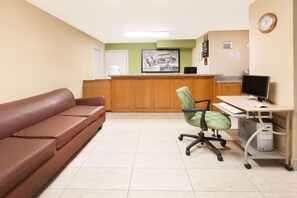 A reception area with a brown sofa, a green office chair, a desk with a computer, and a clock on the wall. - Arya Inn & Extended Stay (Longview)