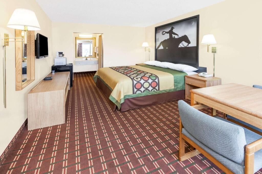 Quarto de hotel com cama, escrivaninha, cadeira, televisão e um quadro na parede.