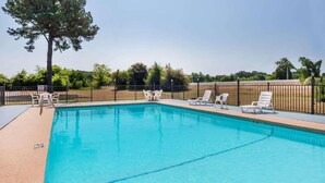 A rectangular swimming pool with clear blue water, surrounded by a black metal fence. There are several white lounge chairs and a single white table near the pool's edge. A large tree is visible to the left of the pool.