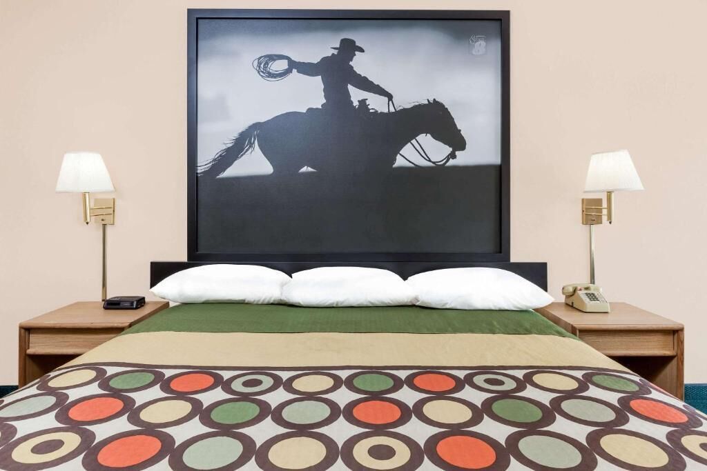 A hotel room with a bed, two bedside lamps, a mounted picture of a cowboy on horseback, and a patterned bedspread.