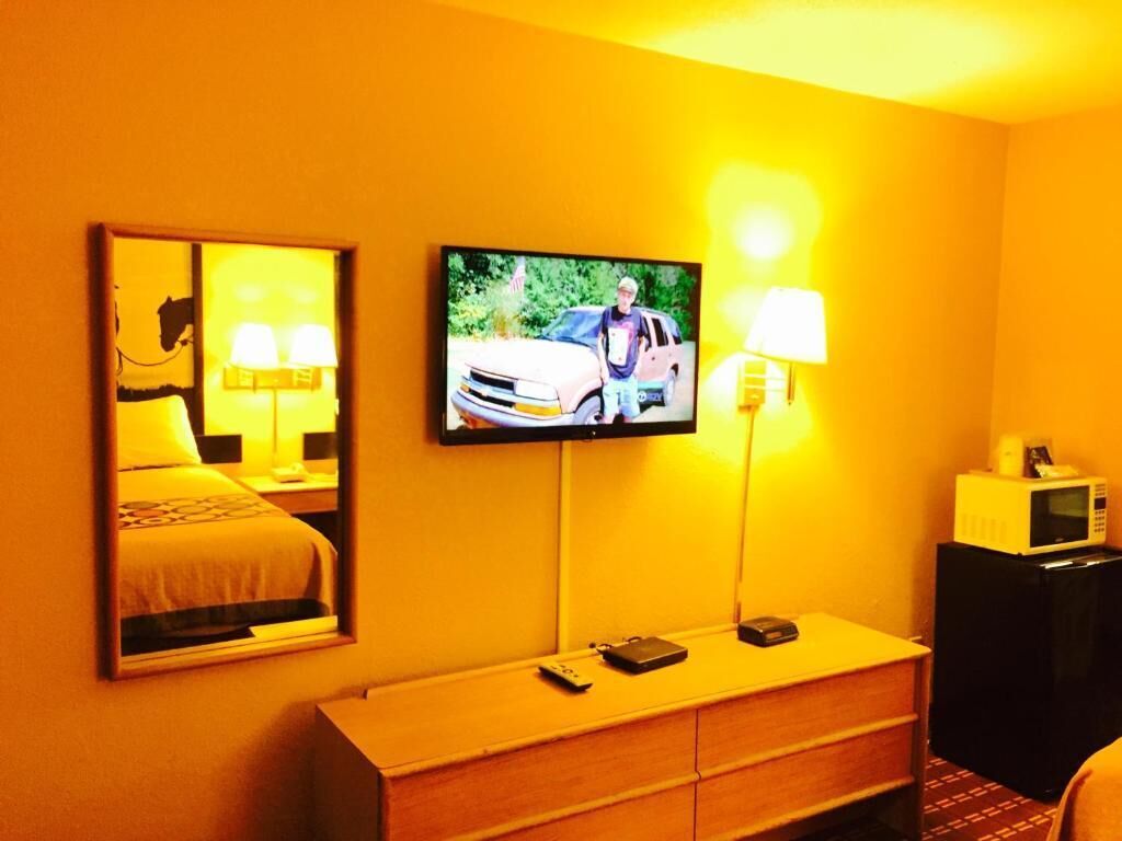 A hotel room with a television mounted on the wall, a bedside table with a lamp, a microwave, and a small cabinet.