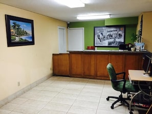 A reception area with a wooden desk, a green office chair, a framed picture on the wall, and a poster. - Arya Inn & Extended Stay (Longview)
