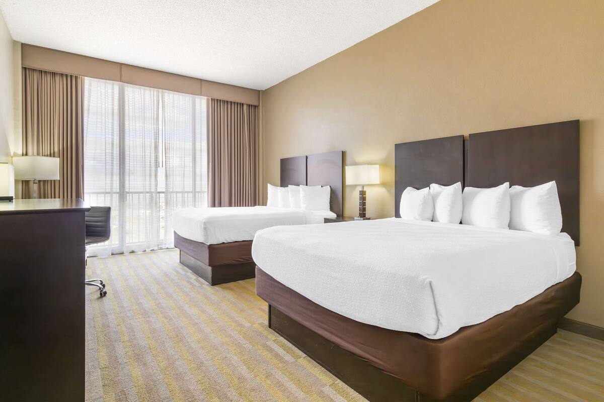 A hotel room with two beds, a desk, and a large window with curtains.