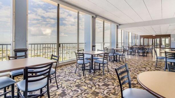 A spacious indoor area with multiple tables and chairs, large windows offering a view of the sky and clouds, and a patterned carpet. - Best Western Corpus Christi (Corpus Christi)