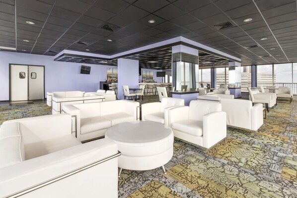 Un moderno vestíbulo de hotel con sofás de cuero blanco, una zona de bar y ventanas amplias que ofrecen vistas de la ciudad.