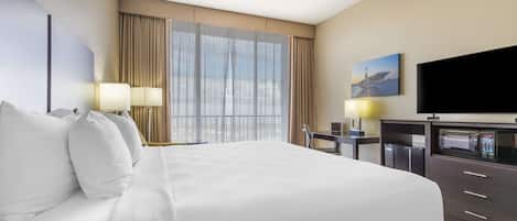 Une chambre d’hôtel avec un grand lit, un bureau avec une chaise, un téléviseur à écran plat, un four à micro-ondes et une grande fenêtre avec des rideaux.