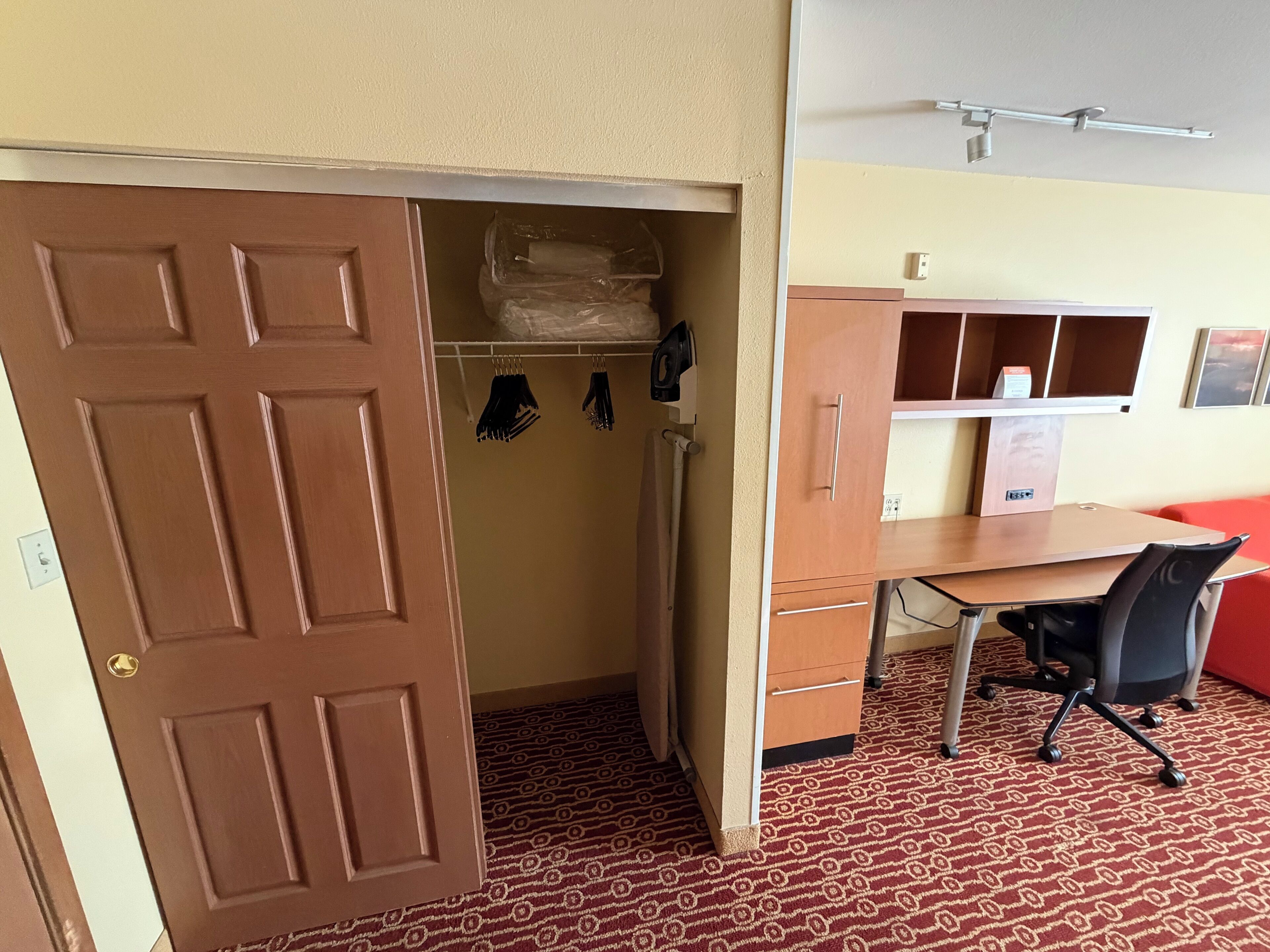 A room with a wooden desk, a chair, a wardrobe, and a door leading to a closet with hanging clothes.
