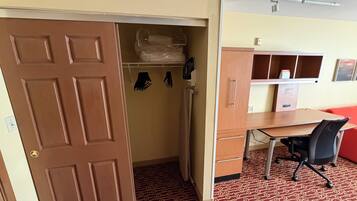 A room with a wooden desk, a chair, a wardrobe, and a door leading to a closet with hanging clothes.
