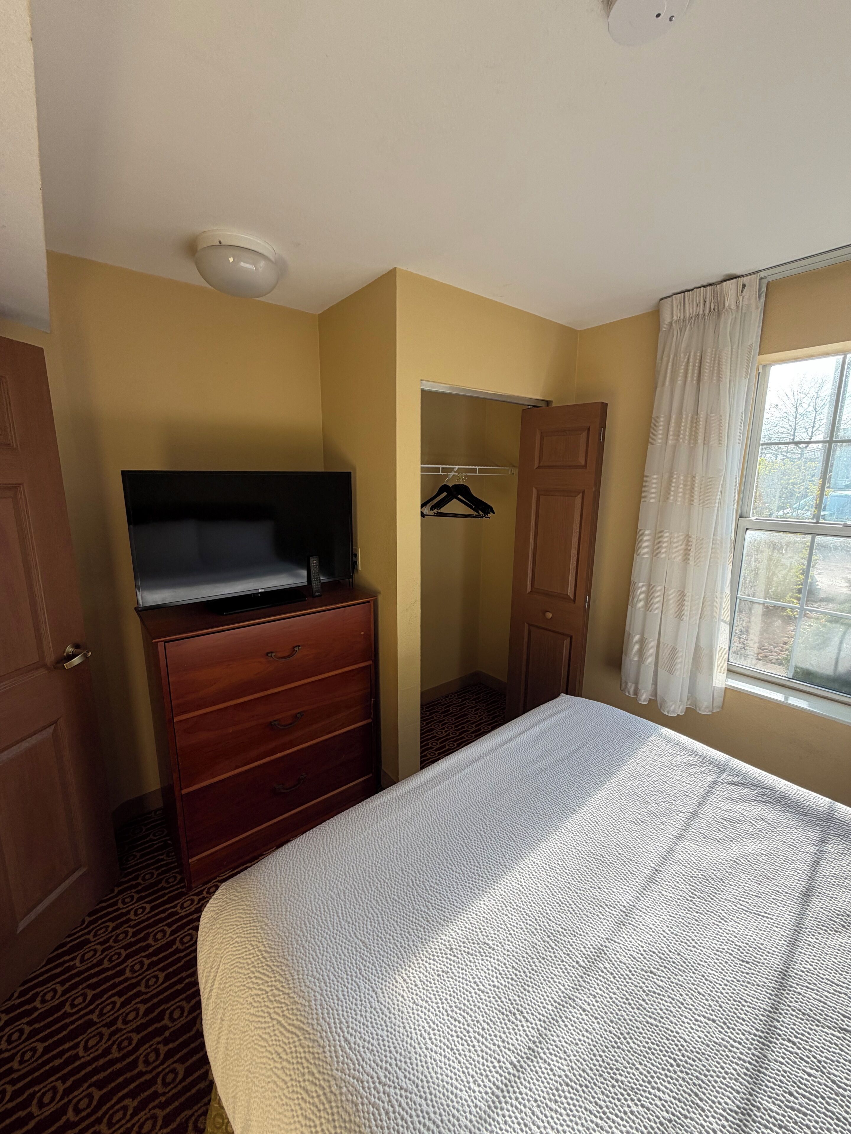 Quarto de hotel com uma cama, penteadeira, televisão, armário e janela com cortinas.