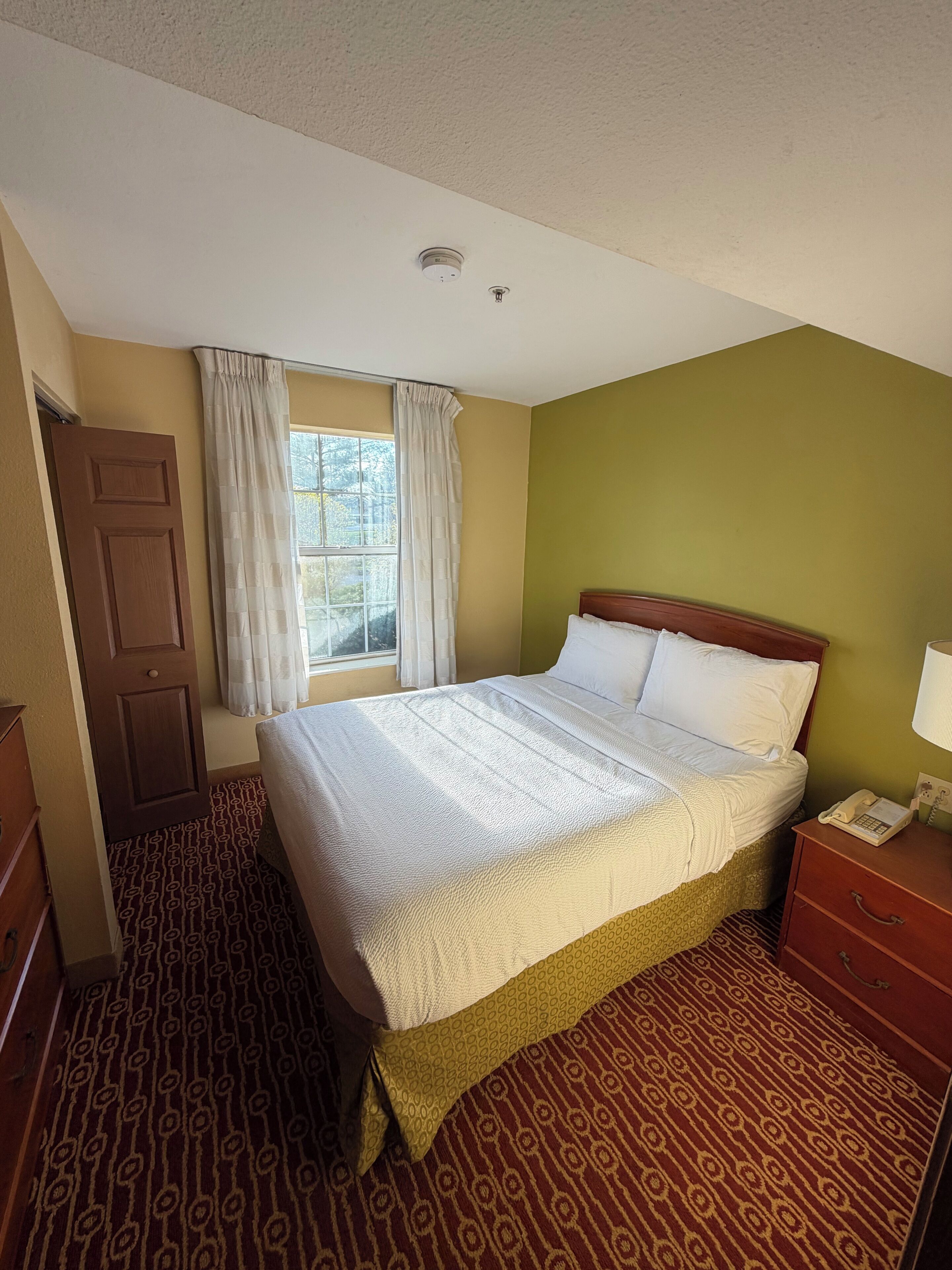 A hotel room with a bed, a bedside table with a lamp, a window with curtains, a door, and a patterned carpet.