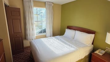 A hotel room with a bed, a bedside table with a lamp, a window with curtains, a door, and a patterned carpet.