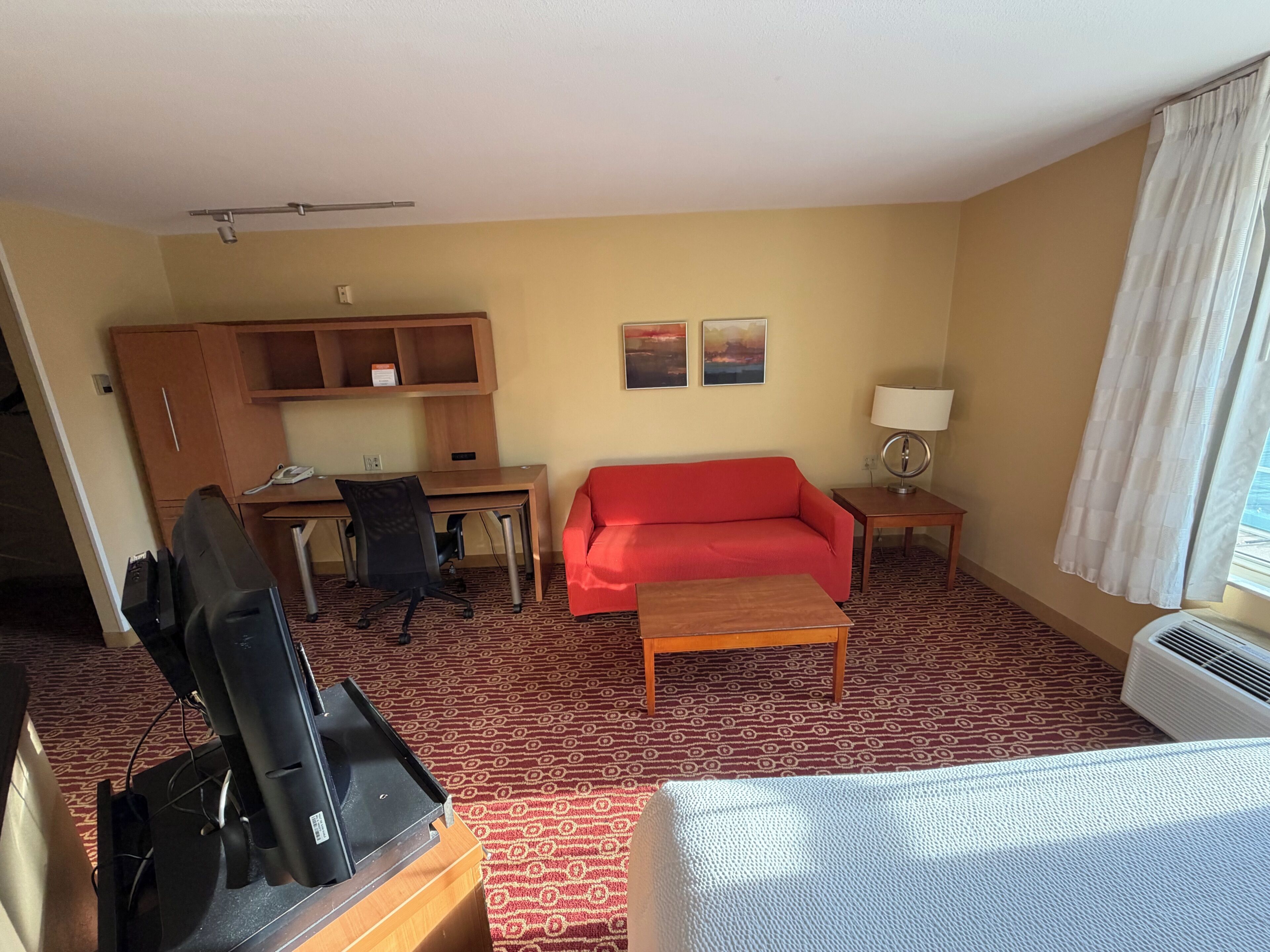 Une chambre d’hôtel avec un bureau, un canapé rouge, une petite table, une lampe, une télévision et un lit.