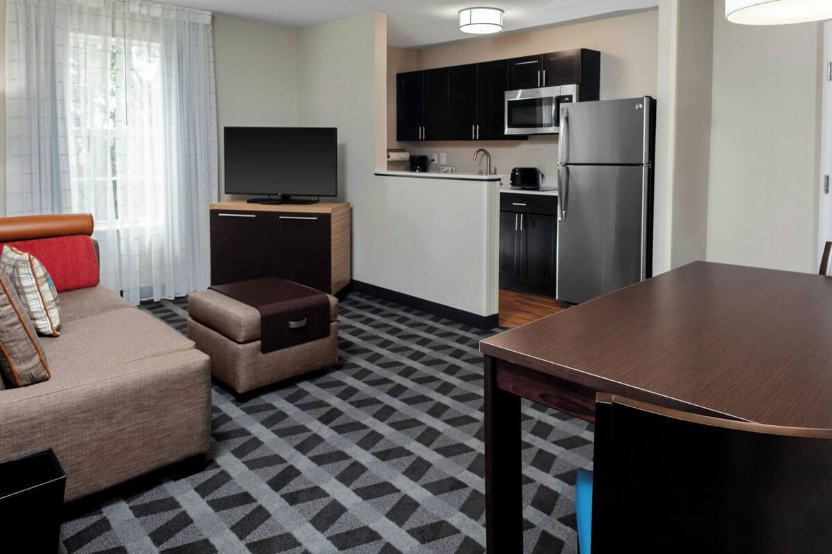 A compact hotel room with a sofa, ottoman, dining table, kitchenette, and a flat-screen TV.