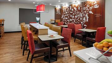 Un comedor moderno con suelo de madera, sillas tapizadas en rojo y marrón, y mesas. Hay una pared decorativa con ilustraciones blancas y una lámpara colgante roja sobre las mesas.