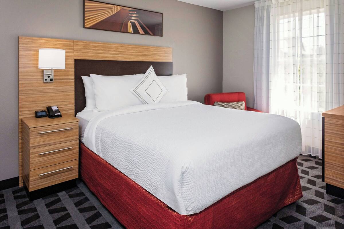 A hotel room with a large bed, a wooden headboard, a bedside table with a lamp, a telephone, and a red chair.