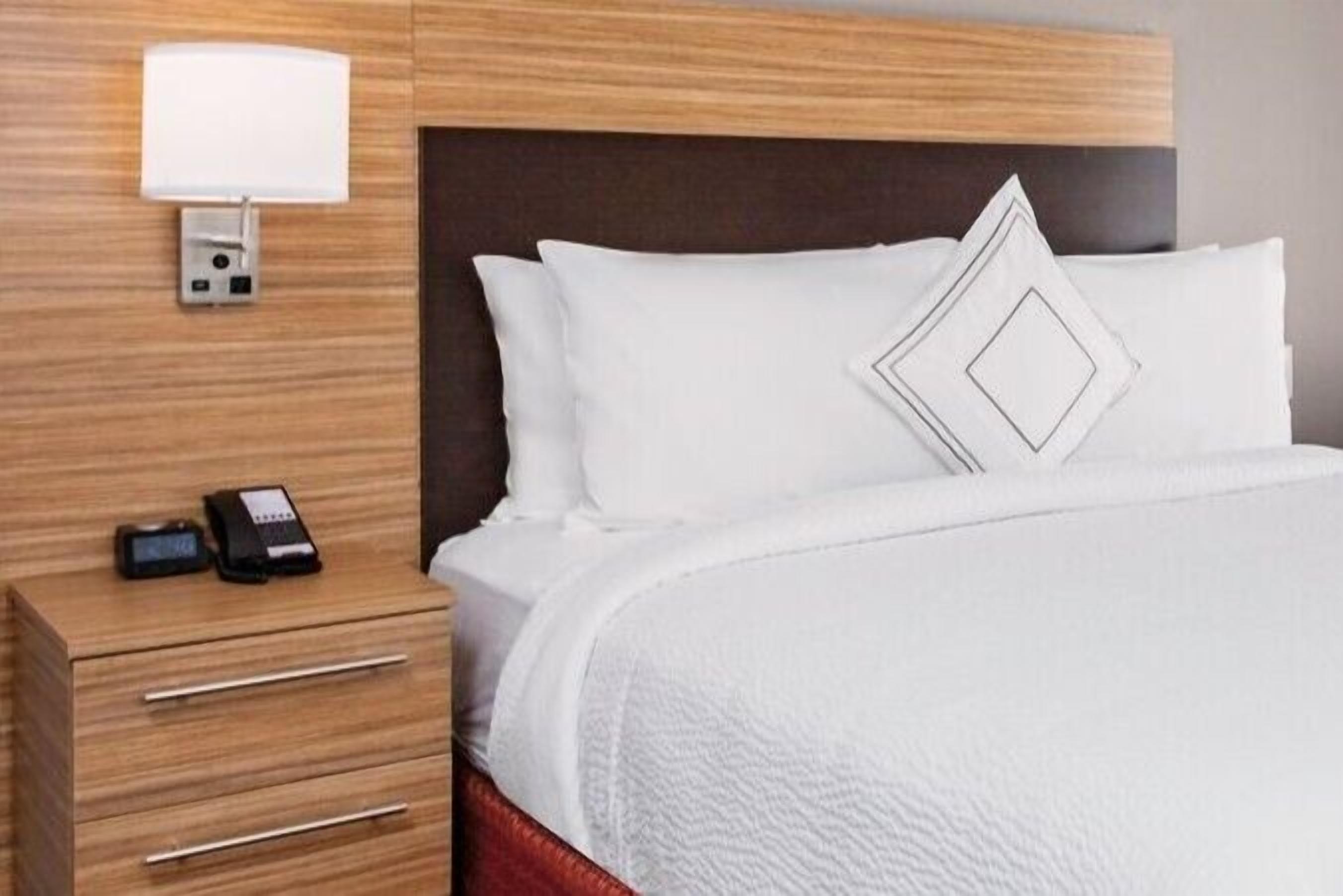 Una camera d'albergo con una testiera in legno, un comodino con una lampada, un telefono e un letto con biancheria bianca e un cuscino geometrico.