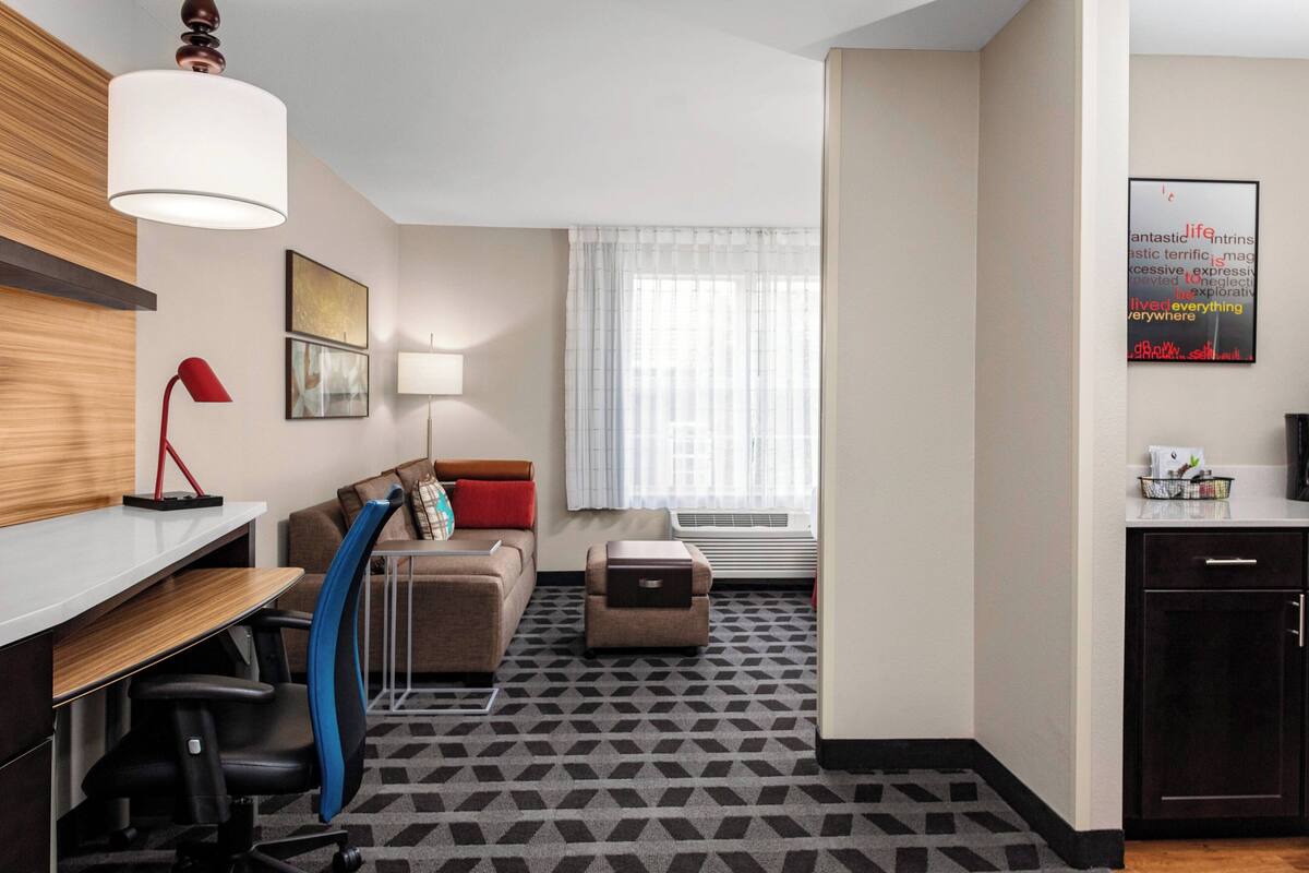 A modern hotel room with a desk, a sofa, a small ottoman, a red lamp, and a window with sheer curtains.