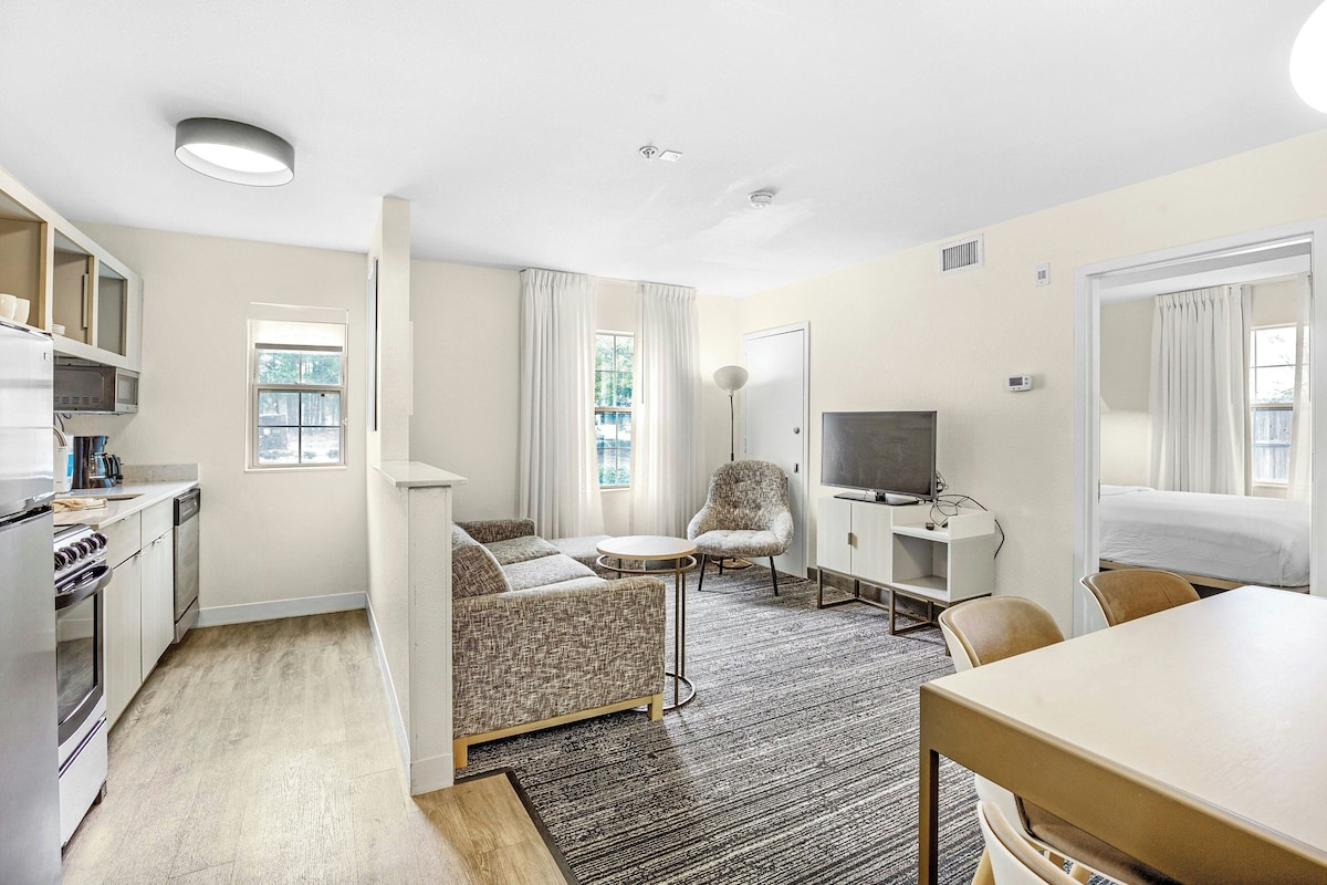 Habitación de hotel compacta con kitchenette, un sofá, una pequeña mesa redonda, un televisor y una cama.
