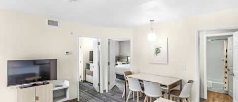 Une chambre d’hôtel moderne dotée d’une télévision à écran plat, d’un coin repas avec une table et des chaises, d’une petite table ronde et d’un espace douche avec une porte en verre.