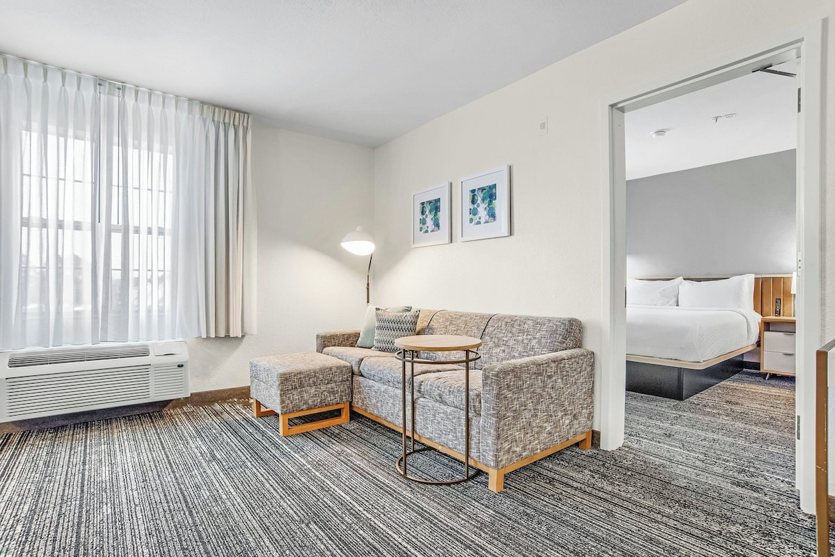 Una habitación de hotel moderna con un sofá gris, una pequeña mesa redonda, una cama con ropa blanca, una mesita de noche y un gran ventanal con cortinas translúcidas.