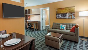 A hotel room with a flat-screen TV, a sofa with a decorative pillow, a dining table set with plates and glasses, and a floor lamp. - TownePlace Suites by Marriott Savannah Midtown (Savannah)