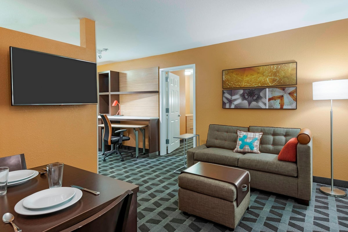 Hotelkamer met een flatscreen televisie, een bank met decoratief kussen, een eettafel gedekt met borden en glazen, en een vloerlamp.