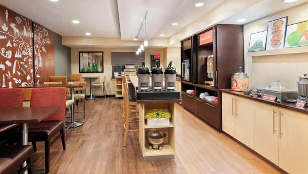 A modern cafe with wooden flooring, a coffee bar, seating area, and a kitchen in the background. - TownePlace Suites by Marriott Savannah Midtown (Savannah)