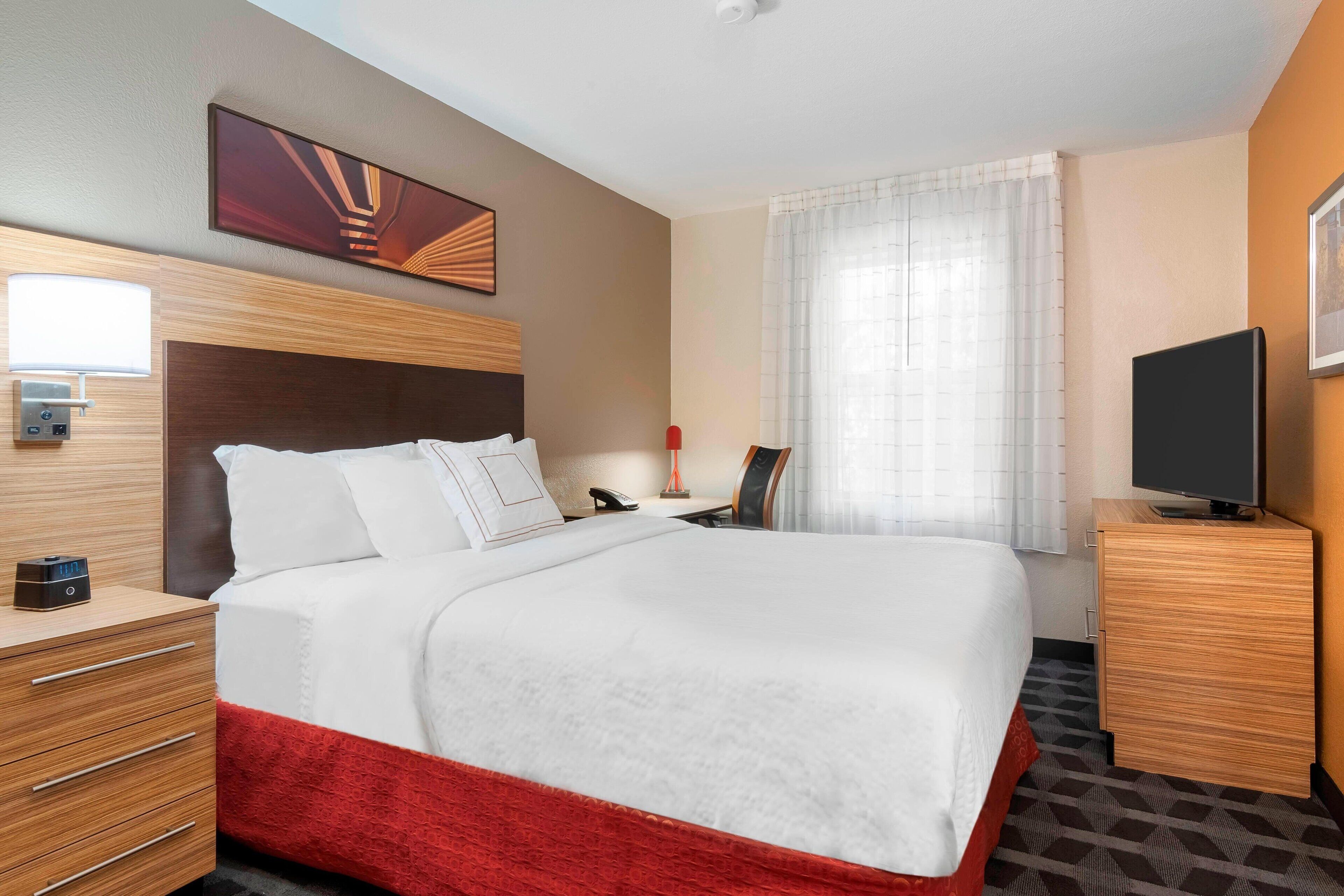 A hotel room with a large bed, wooden headboard, a bedside table with a lamp, a TV on a stand, and a painting on the wall.