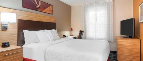 A hotel room with a large bed, wooden headboard, a bedside table with a lamp, a TV on a stand, and a painting on the wall.