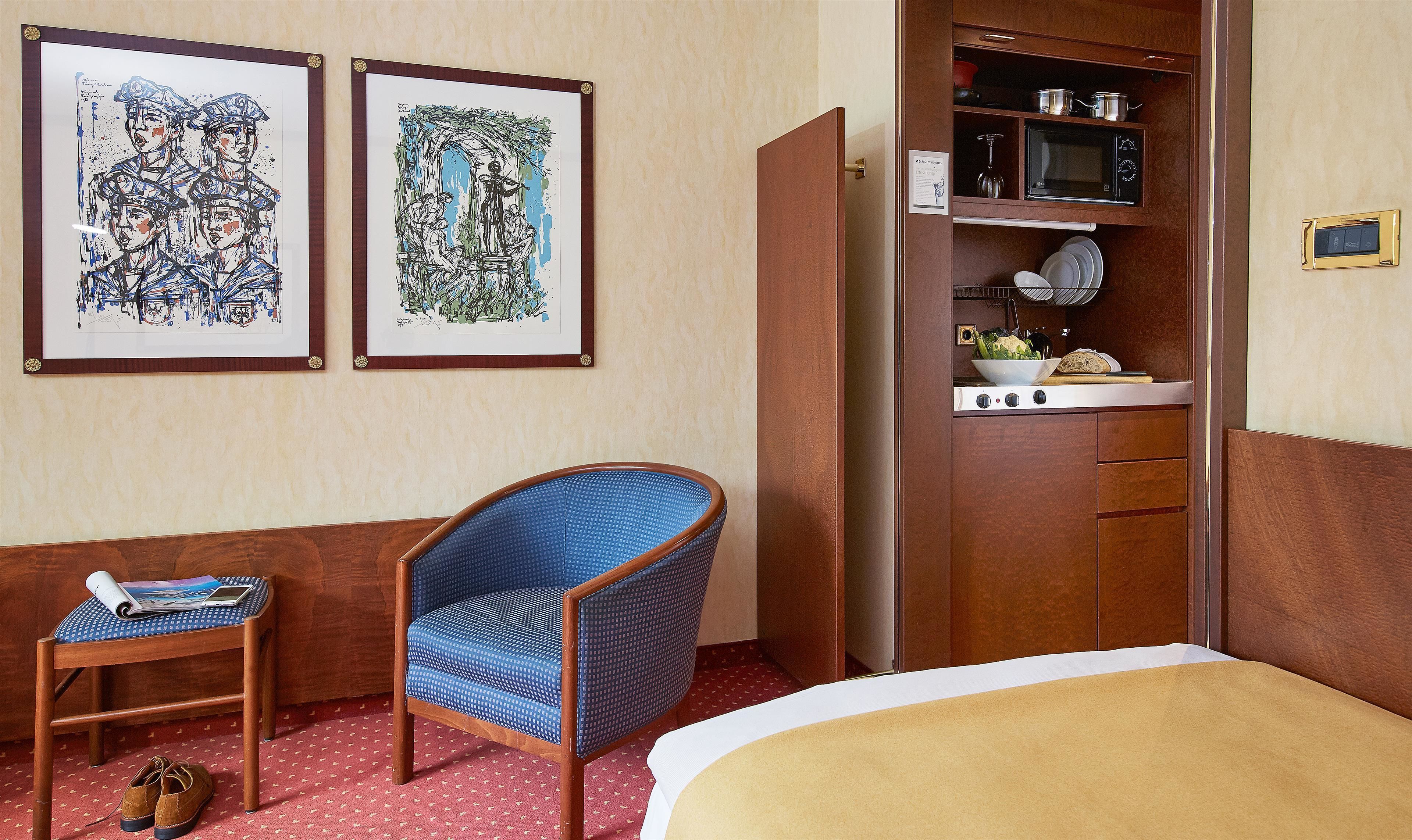 A hotel room with a bed, a chair, a small table with a magazine, and two framed artworks on the wall.