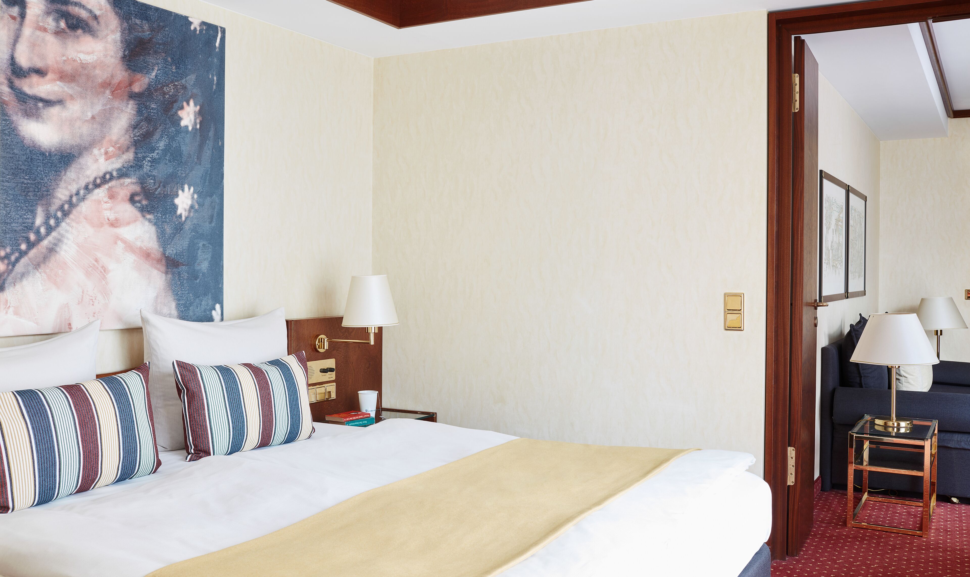 A hotel room with a large bed, a nightstand, a lamp, a painting on the wall, and a view of another room.