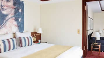 A hotel room with a large bed, a nightstand, a lamp, a painting on the wall, and a view of another room.
