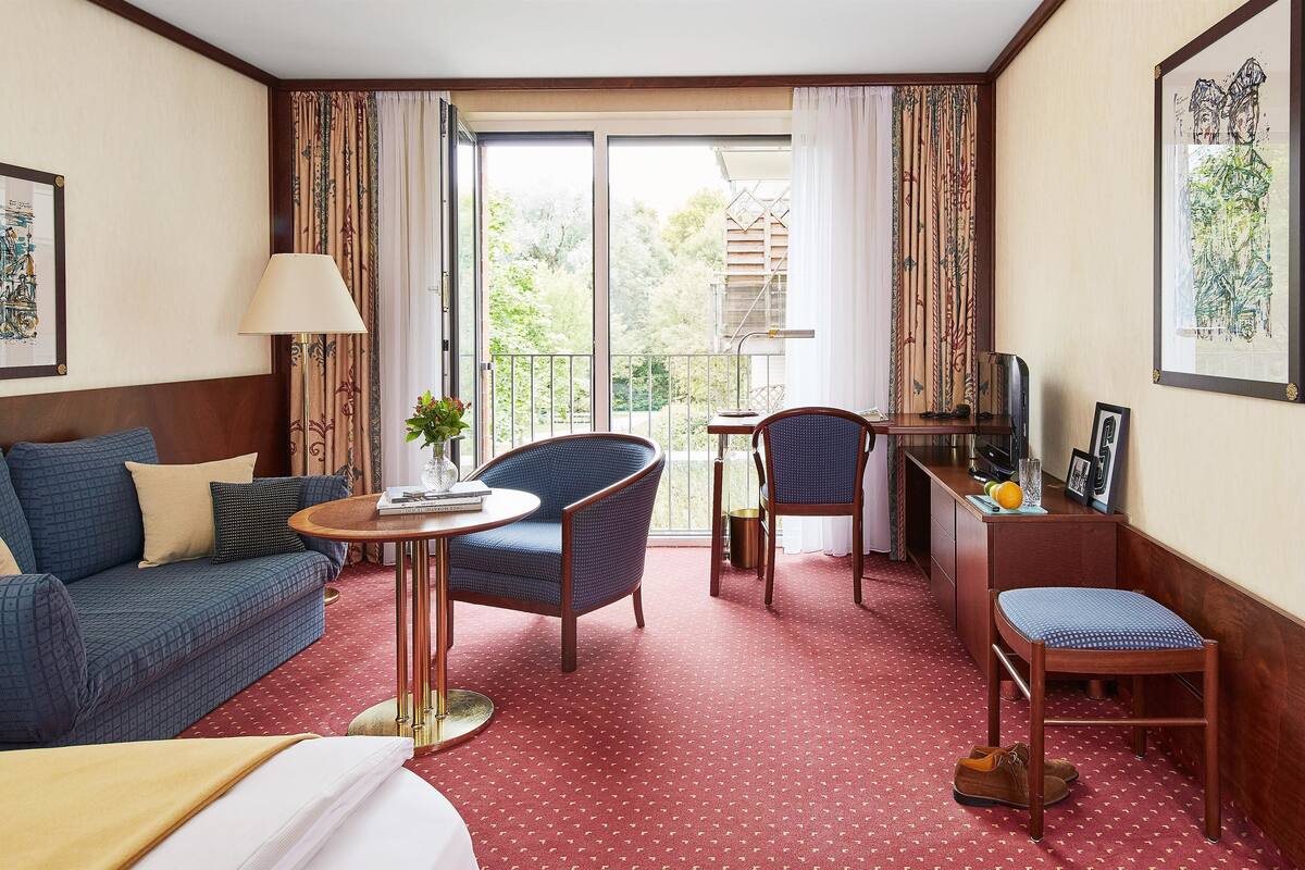 A hotel room with a sofa, a chair, a small table, a desk with a computer, and a balcony with a view of greenery.