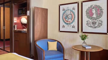 Una habitación de hotel con una silla azul estampada, una mesita redonda con un florero y dos cuadros en la pared.