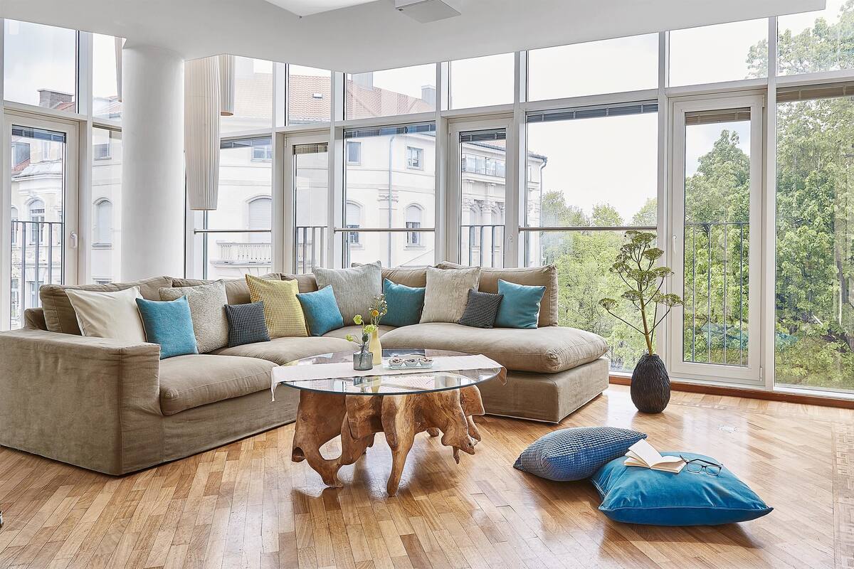 A modern living room with a beige sectional sofa, a round wooden coffee table, and a large window offering a view of buildings and trees.