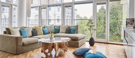 Een moderne woonkamer met een beige hoekbank, een ronde houten salontafel en een groot raam met uitzicht op gebouwen en bomen.