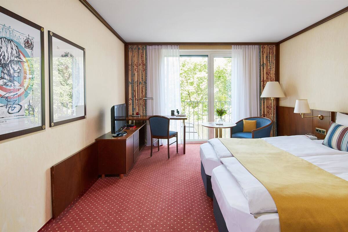 A hotel room with a bed, a desk, a chair, a small table, and a balcony with curtains.