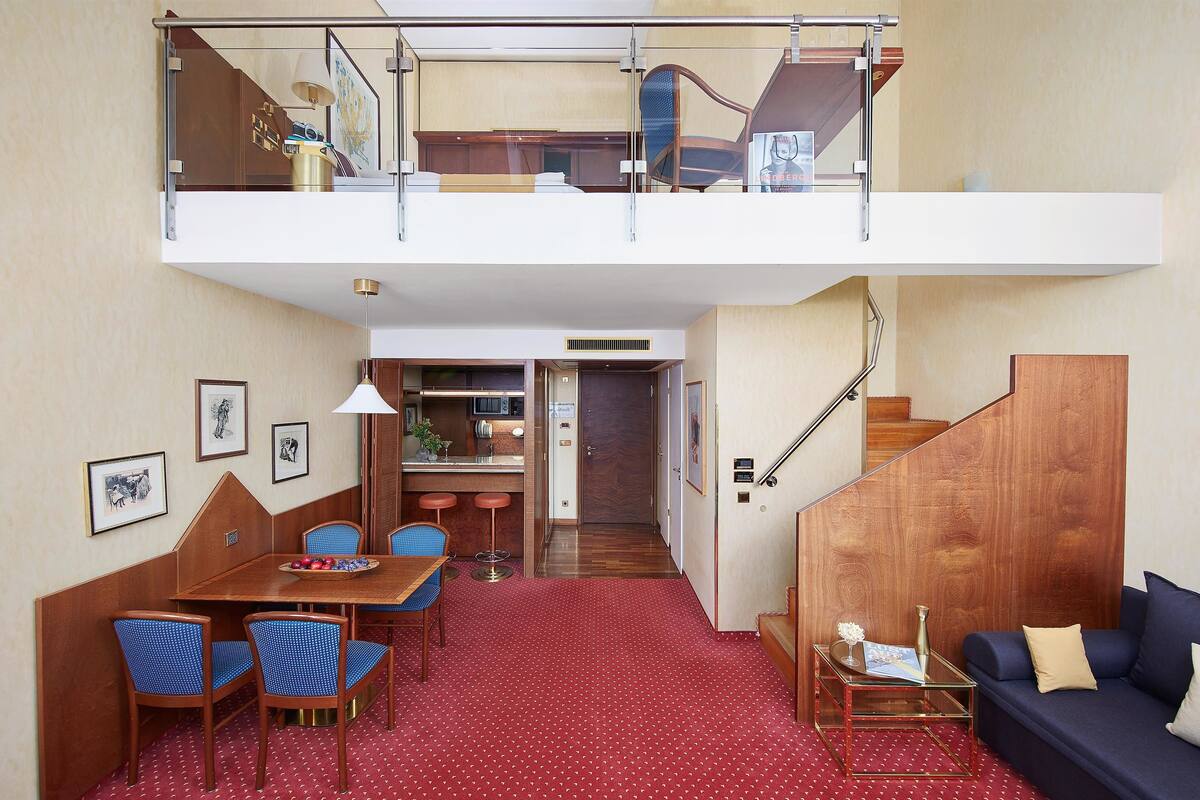 A compact hotel room with a dining area, a sofa, and a staircase leading to an upper level.