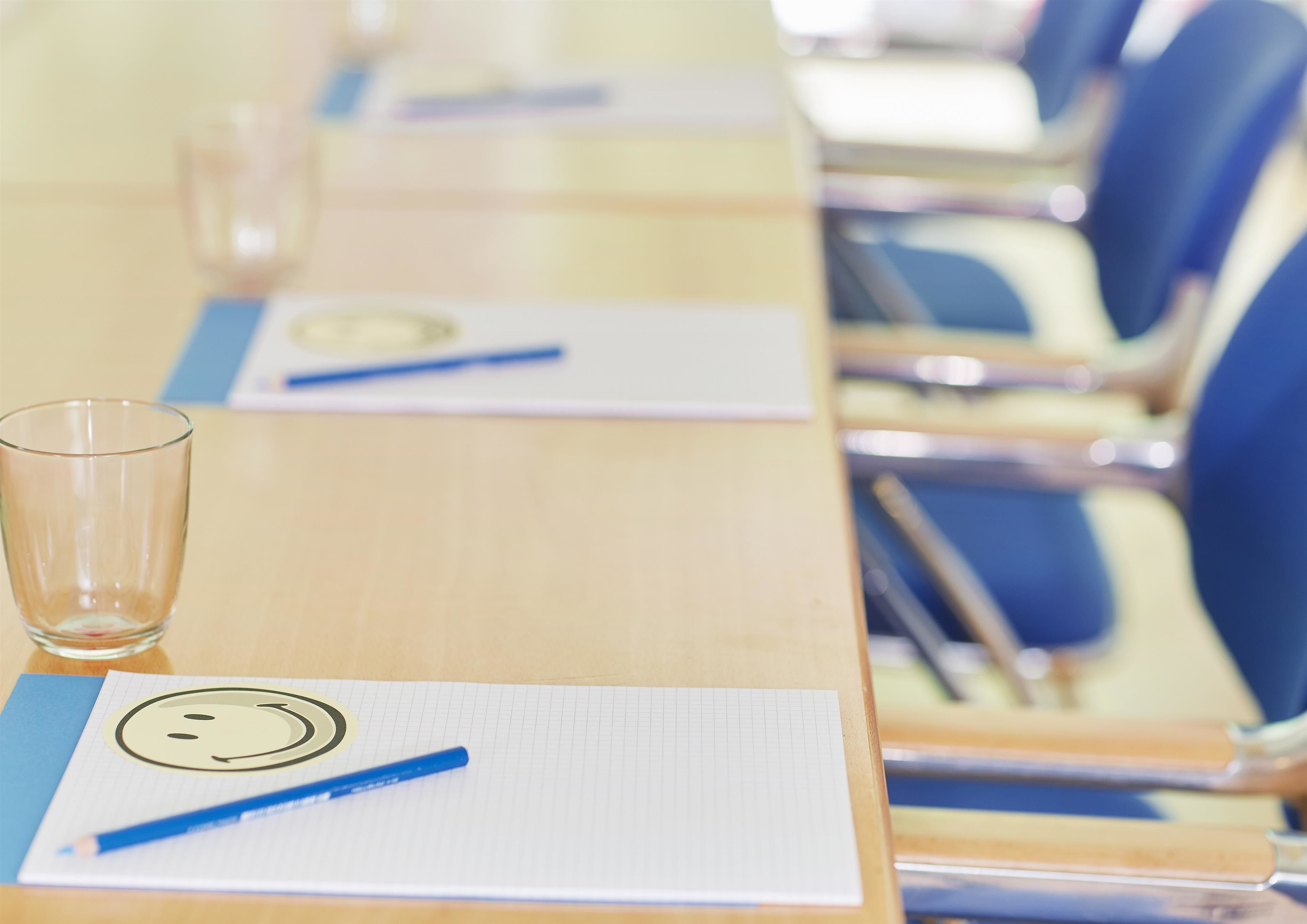 Una mesa de conferencias con un vaso, un bloc de notas con una carita sonriente y un bolígrafo azul.