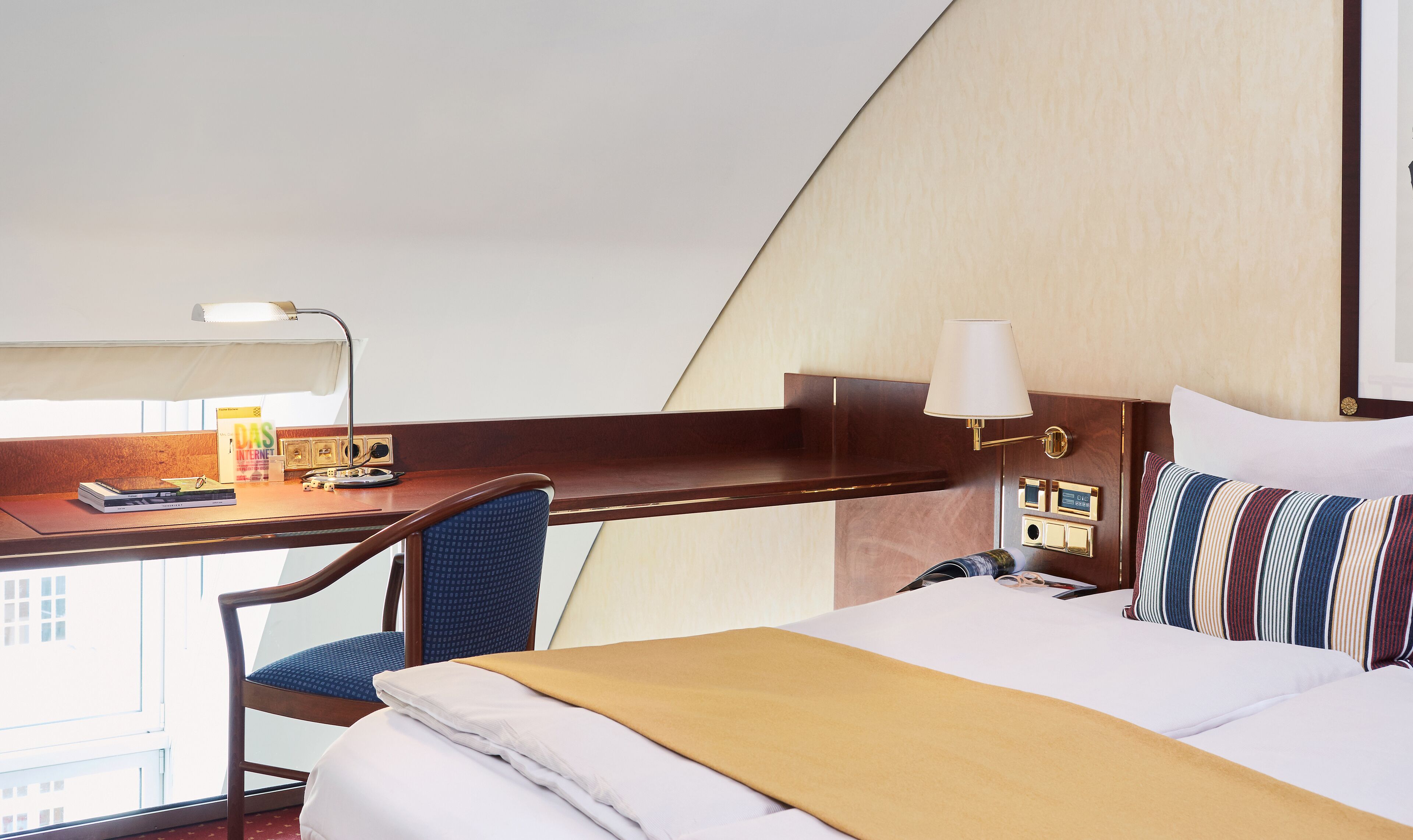 A hotel room with a bed, a desk, a chair, a lamp, and a window.