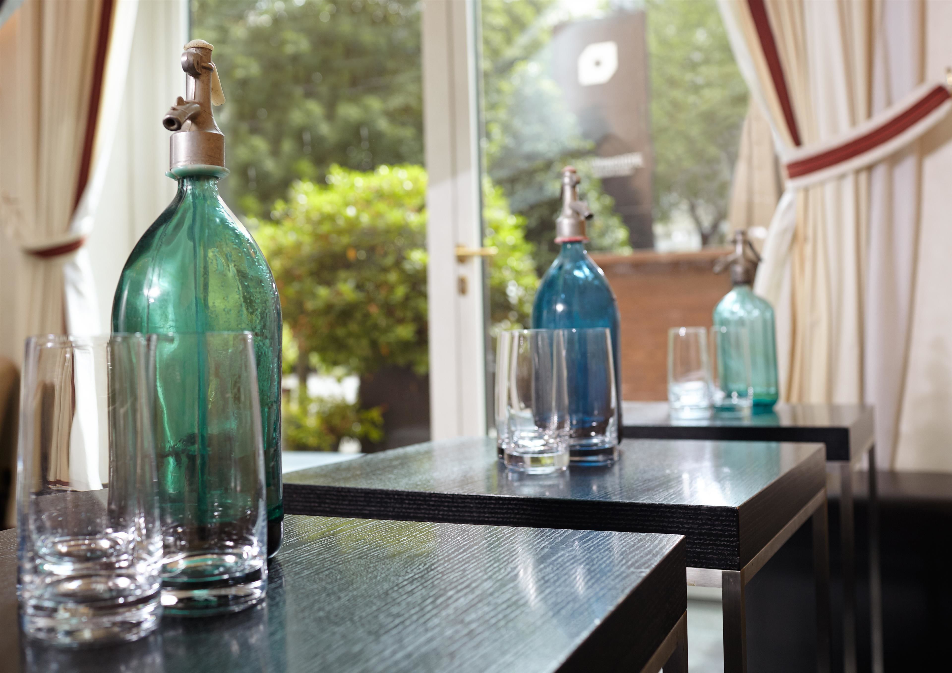 A table with glassware and bottles, a window with curtains, and a view of greenery outside.