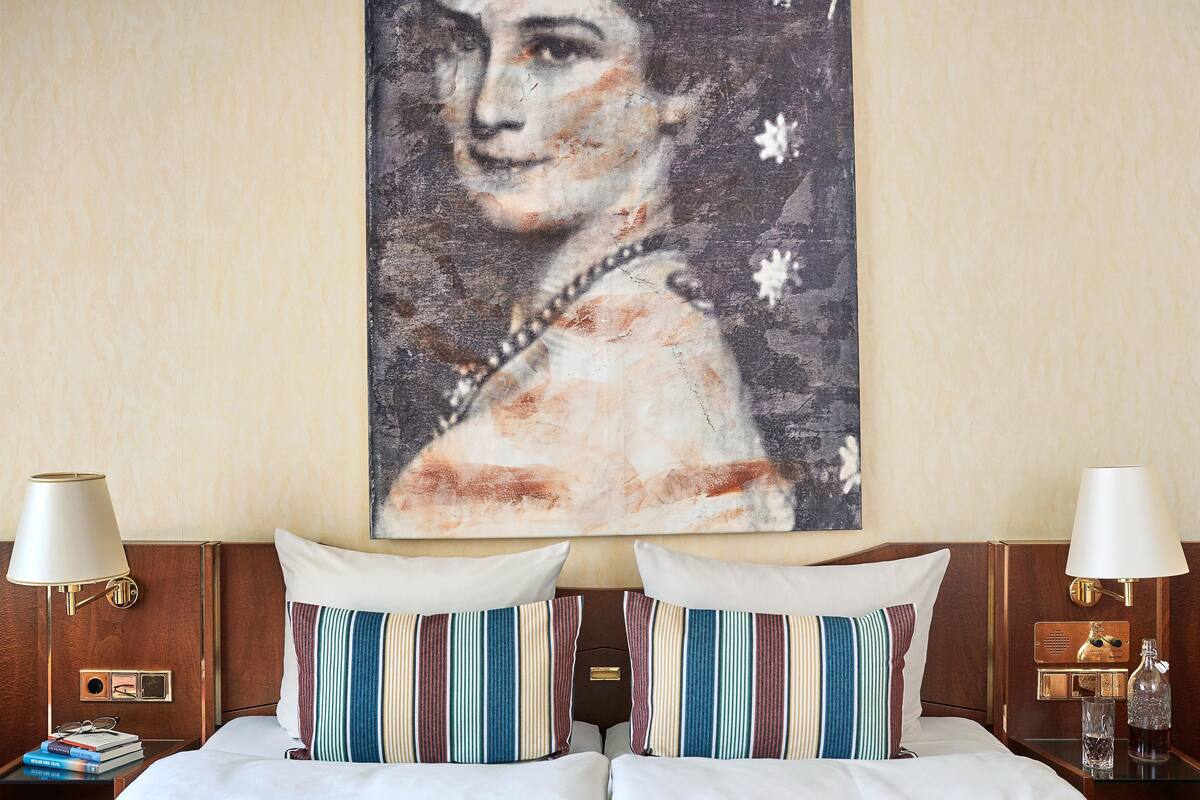 A hotel room with a bed, striped pillows, a painting on the wall, and two bedside tables with lamps and a bottle.