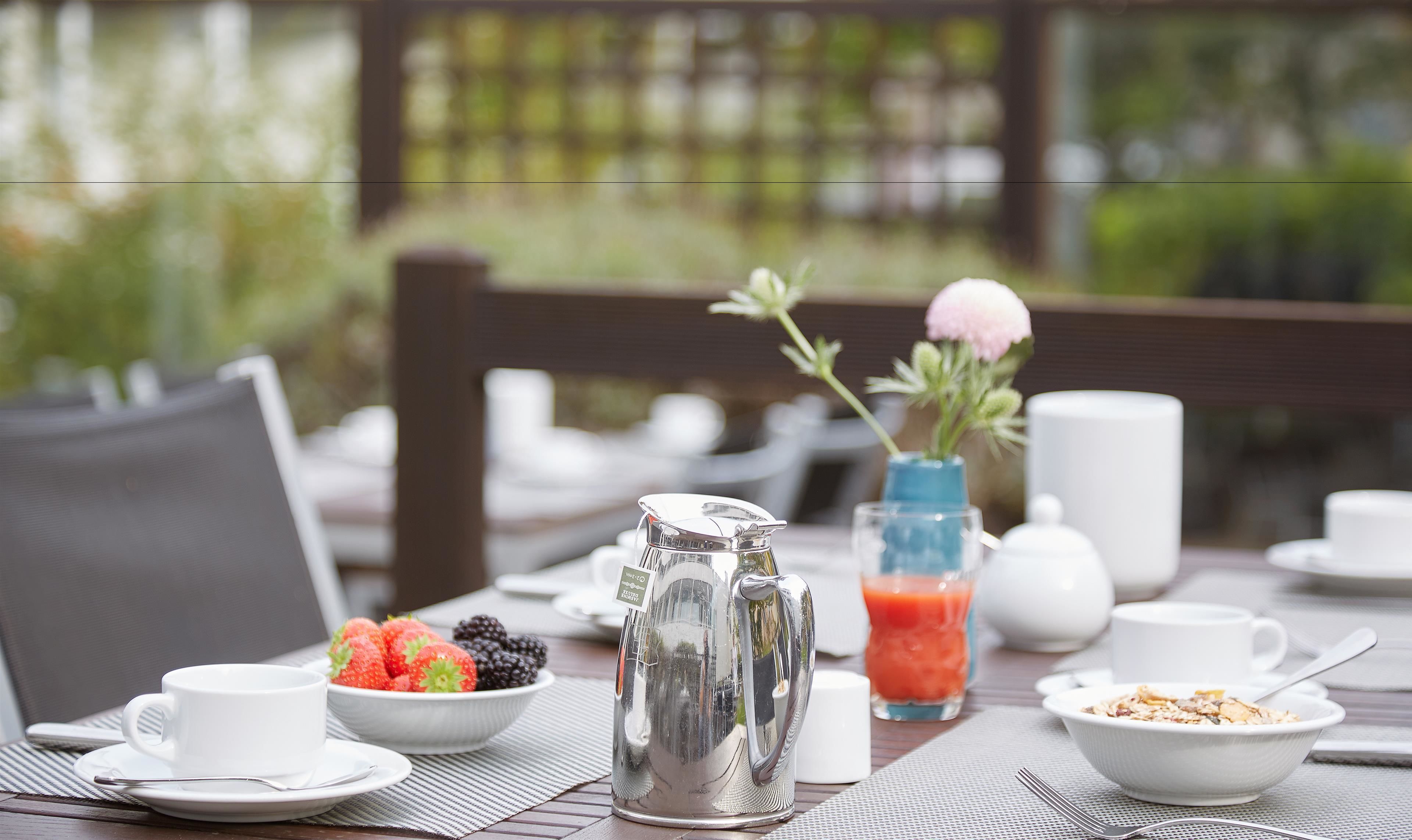 A breakfast table with a pitcher, a bowl of fruit, a glass of juice, and a vase with flowers.