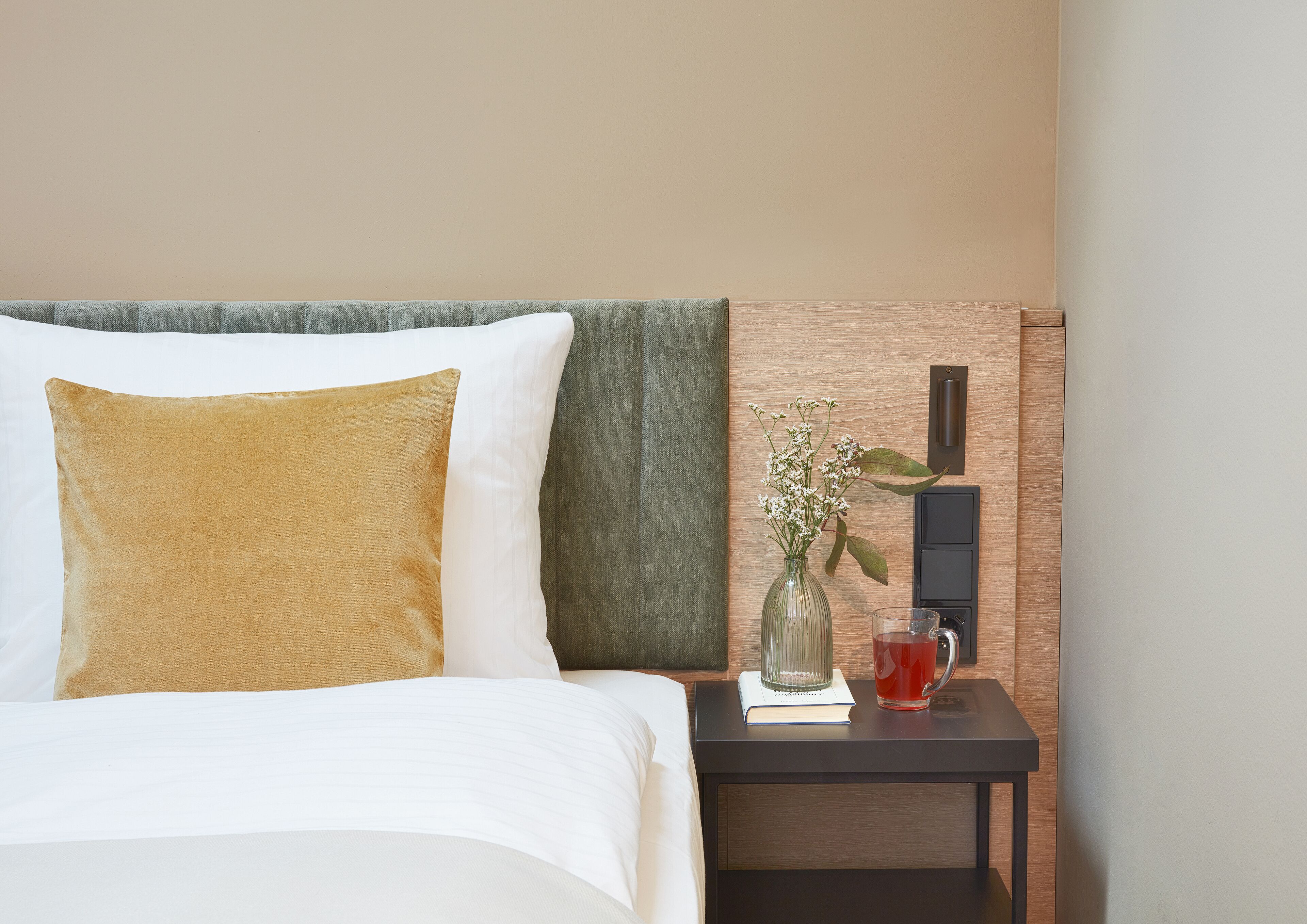 Una cama bien hecha con un cojín blanco y dorado, un cojín verde y una mesita de noche con un florero de flores y un vaso de bebida roja.